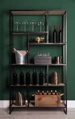 Rack with bottles of wine and glasses near green wall