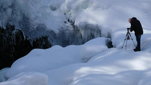 Old bold photographer in the snow with tripod and camera takes pictures of the waterfall.