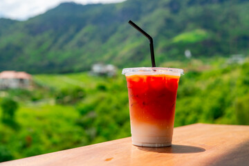 Iced milk tea with ice in a plastic cup Laying on a wooden table behind is a view of the mountains and the bright sky.
