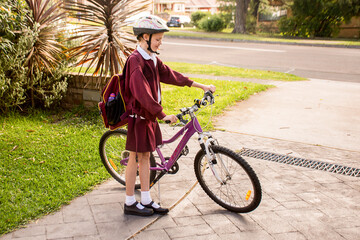 A girl wearing maroon school unfiform and bike helmet going to school on her bicycle. School students return to classrooms after COVID-19 outbreak
