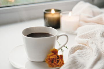 fall morning cup of coffee and orange flower. hygge concept, warm knitted blanket and burning candles on background. cozy autumn home decor.
