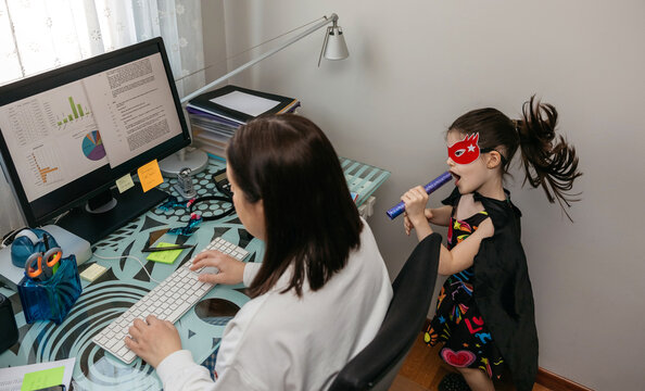 Top View Of Woman Working From Home With Her Daughter Singing And Playing By Her Side. Selective Focus On Girl In Background