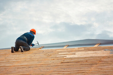 Worker hammer nails in wooden board on roof at height. Roofer carpenter works on roof in hardhat and safety harness. Renovation, repair and fixing frame work house. New building construction