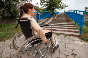 Fototapeta na wymiar A young disabled girl sits in a wheelchair in front of the stairs. The concept of a wheelchair, disabled person, full life, paralyzed, disabled person, health care, loneliness.