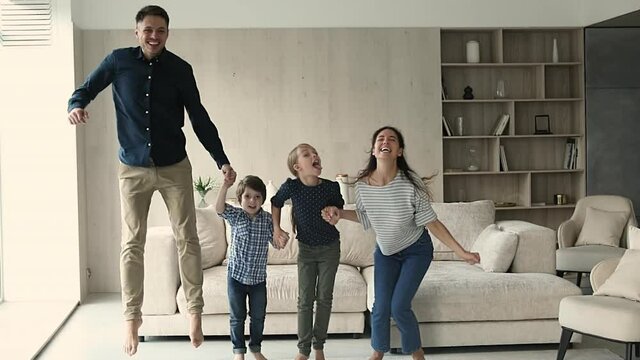 Overjoyed candid happy young couple parents holding hands of laughing little kids son and daughter, jumping together in modern living room in slow motion, rental tenancy dwelling ownership concept.