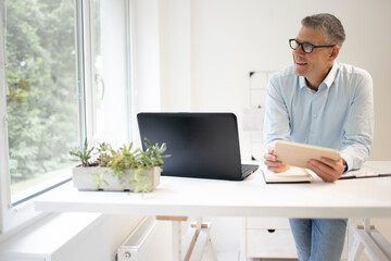 business man with blue shirt and black glasses is standing behind standing table and is working with his tablet and a black laptop in a modern office