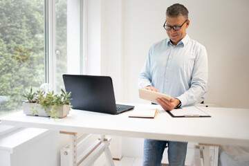 business man with blue shirt and black glasses is standing behind standing table and is working with his tablet and a black laptop in a modern office