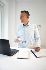 business man with blue shirt and black glasses is standing behind standing table and is working with his tablet and a black laptop in a modern office