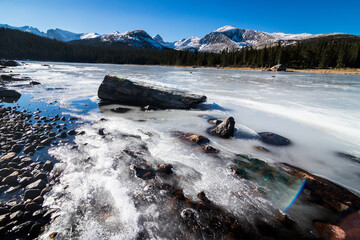 A wide angle landscape shot of a frozen lake with a log and rocks in it with mountains and a pine tree line in the background in winter in Colorado