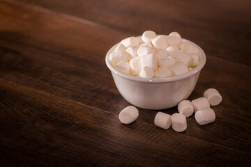 Fototapeta na wymiar A close up shot of a bowl of small, white marshmallows with marshmallows fallen on the wooden table, studio shot of marshmallow bowl on kitchen table