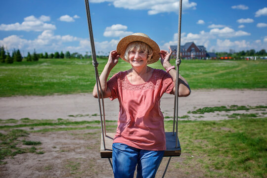 Carefree senior woman in straw hat swinging on a wooden swing and looking at the camera, mental and physical health, age beauty, baby boomer generation, summer lifestyle