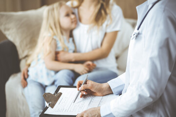 Fototapeta premium Doctor and patient. Pediatrician using clipboard while examining little girl with her mother at home. Sick and unhappy child at medical exam. Medicine concept
