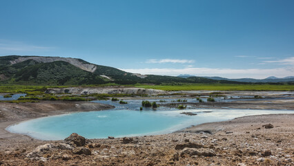 An amazing turquoise geothermal lake in the caldera of an extinct volcano. There is almost no vegetation on the stony soil. A mountain range against the blue sky. A summer day. Kamchatka. Uzon