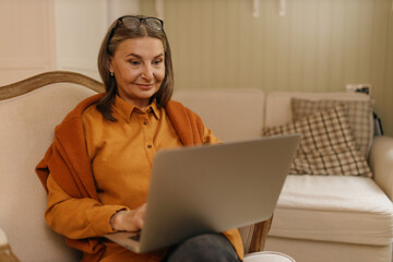 Attractive confident female on retirement sitting on comfortable armchair with portable computer on...