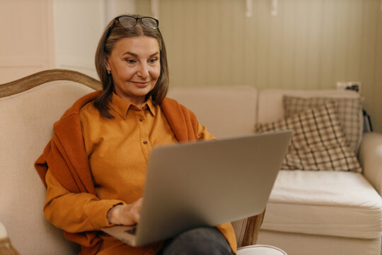 Attractive confident female on retirement sitting on comfortable armchair with portable computer on her lap, surfing internet, shopping online, enjoying wireless high speed connection at home