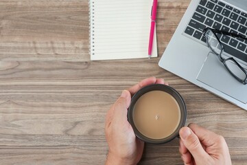 Cropped hand holding a cup of coffee, a laptop, and empty notebook on the wooden table