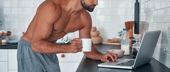 Handsome young shirtless man using laptop while drinking tea at home kitchen