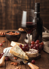 Glass and bottle of red wine with selection of various cheese on the board and grapes on wooden background. Blue Stilton, Red Leicester and Brie Cheese and bowl of nuts.