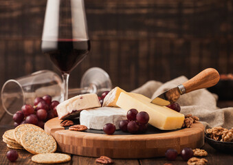 Glass of red wine with selection of various cheese on the board and grapes on wooden background. Blue Stilton, Red Leicester and Brie Cheese and bowl of nuts and crackers.