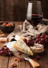 Glass of red wine with selection of various cheese on the board and grapes on wooden background. Blue Stilton, Red Leicester and Brie Cheese and bowl of nuts.