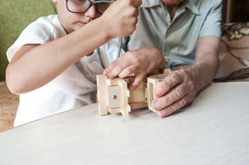Grandfather and grandson assemble a wooden toy car at home using screwdriver tools. Relations between generations