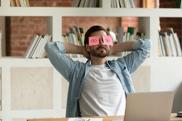Male employee puts hands behind head lean on chair relaxing seated at workplace desk with pink sticky notes on his eyes. Work accomplishment, unmotivated lazy office clerical worker take break concept - obrazy, fototapety, plakaty