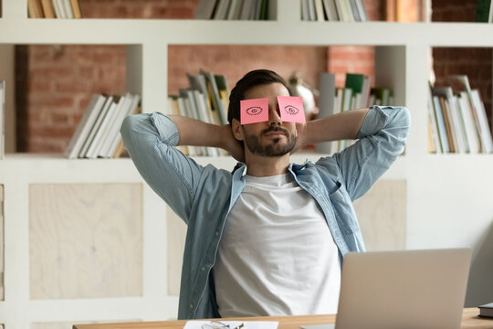 Male employee puts hands behind head lean on chair relaxing seated at workplace desk with pink sticky notes on his eyes. Work accomplishment, unmotivated lazy office clerical worker take break concept