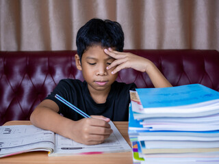 boy doing homework and reading on a wooden table with a pile of books beside The background is a red sofa and cream curtains.