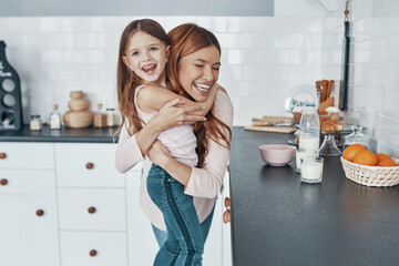 Young beautiful loving mother embracing her daughter and smiling while preparing something at the...