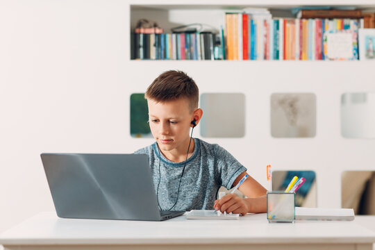 Young little schoolar boy in headset sitting at table with laptop and preparing to school. Online education concept