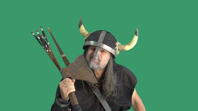 Emotional viking mature man in helmet with horns shaves with battle ax and threatens his enemies. Green background