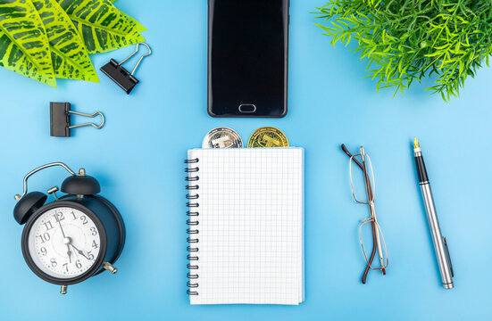 Business still life with crypto currency coins in a blank notebook , alarm clock, mobile phone, glasses, pen and paper clips on blue with fresh green plants to the corners