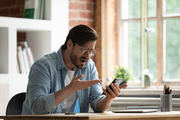 Man sit at desk holds gadget experiences problem with cell phone looks at screen feels annoyed. Bad news, spam in mailbox, stuck or discharged device need repair, wifi connection lost, malware concept