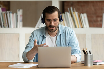 Obraz premium Serious man in glasses headphones sit at workplace desk talks through videoconference, take part in online stream communication use laptop, lead business negotiations remotely. Videocall event concept