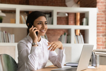 Attractive pleased Indian ethnicity business lady sit at desk with laptop holds smart phone lead...