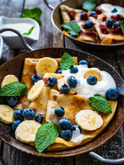 Sweet crepes with bananas, strawberries jam and blueberries in pan on wooden table
