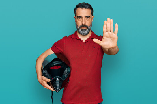 Middle aged man with beard holding motorcycle helmet with open hand doing stop sign with serious and confident expression, defense gesture