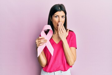 Young hispanic woman holding pink cancer ribbon covering mouth with hand, shocked and afraid for...