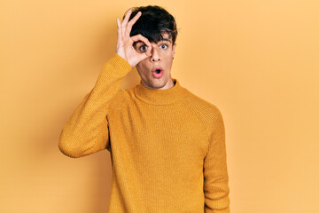 Handsome hipster young man wearing casual yellow sweater doing ok gesture shocked with surprised face, eye looking through fingers. unbelieving expression.