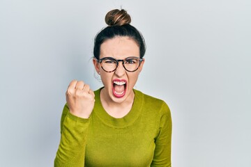 Young hispanic girl wearing casual clothes and glasses angry and mad raising fist frustrated and furious while shouting with anger. rage and aggressive concept.