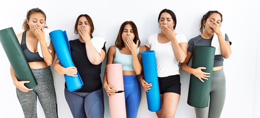 Fototapeta na wymiar Group of women holding yoga mat standing over isolated background bored yawning tired covering mouth with hand. restless and sleepiness.