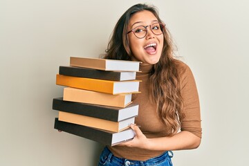 Young hispanic girl holding a pile of books celebrating crazy and amazed for success with open eyes...