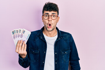 Young hispanic man holding egyptian pounds banknotes scared and amazed with open mouth for surprise, disbelief face - obrazy, fototapety, plakaty