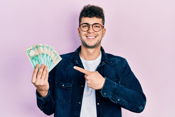 Young hispanic man holding united arab emirates dirham banknotes smiling happy pointing with hand and finger