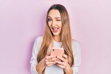 Young blonde girl using smartphone typing message winking looking at the camera with sexy...