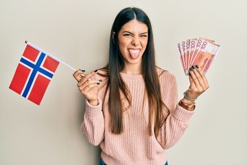 Young hispanic woman holding norway flag and krone banknotes sticking tongue out happy with funny...