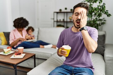 Hispanic father of interracial family drinking a cup coffee yawning tired covering half face, eye...