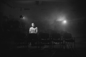 Grayscale shot of a Caucasian man sitting alone in an audience room with studio lights on him