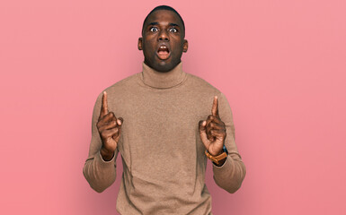 Young african american man wearing casual winter sweater amazed and surprised looking up and pointing with fingers and raised arms.