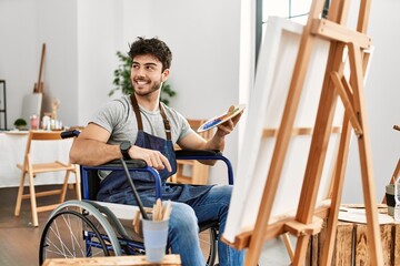 Young hispanic man sitting on wheelchair painting at art studio looking away to side with smile on...